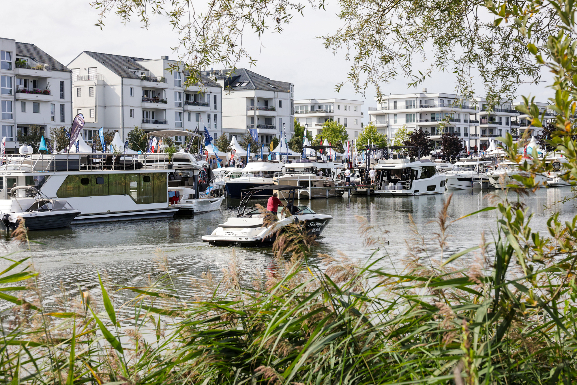 In der schilfumwachsenden Marina Havelauen liegen Boote aller Segmente und Größen an den Stegen. Im Hintergrund sieht man Pagoden an der Hafenpromenade. Ein Motorboot fährt in den Marinahafen ein.