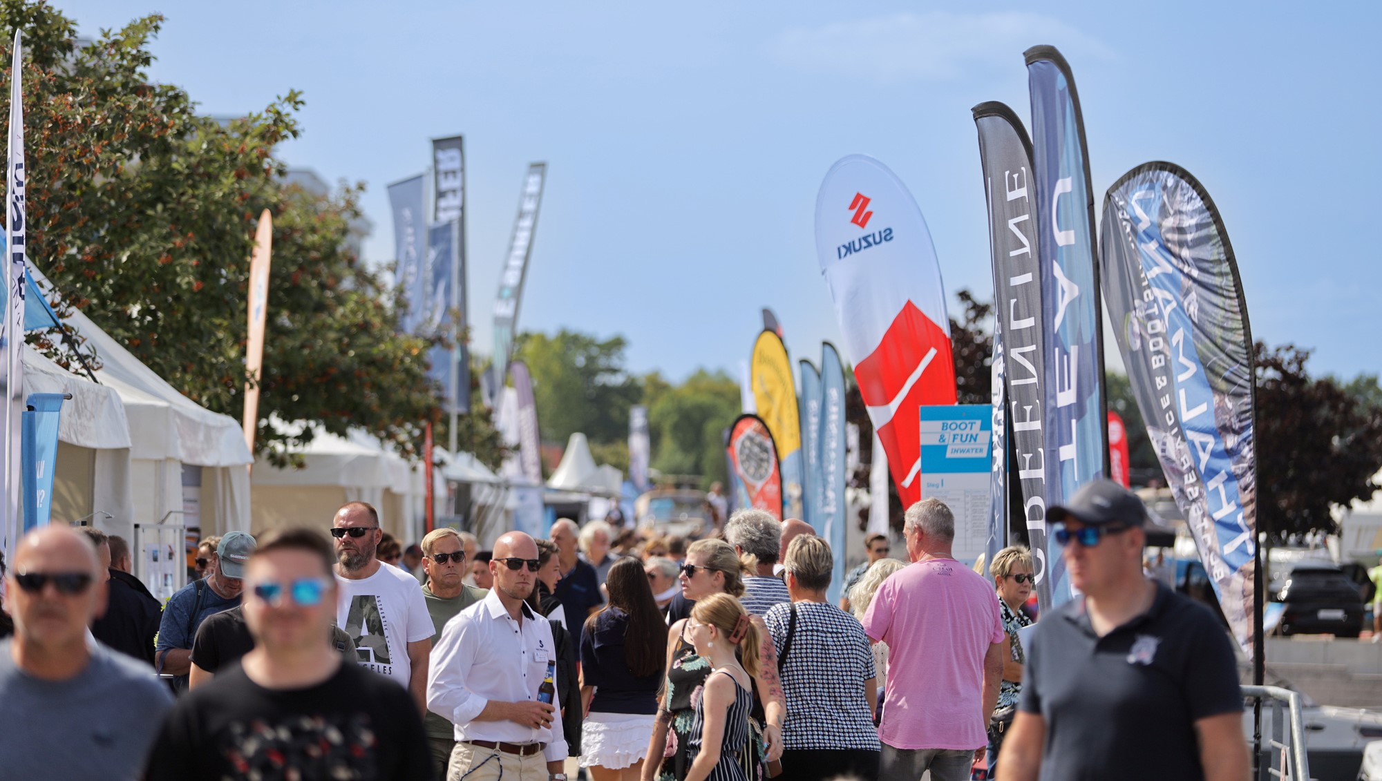 Zwischen Verkaufspagoden, Beachflags und Booten an Stegen: Zahlreiche Besucherinnen und Besucher der BOOT & FUN INWATER 2024 schlendern bei bestem Wetter über die Hafenpromenade der Marina Havelauen.