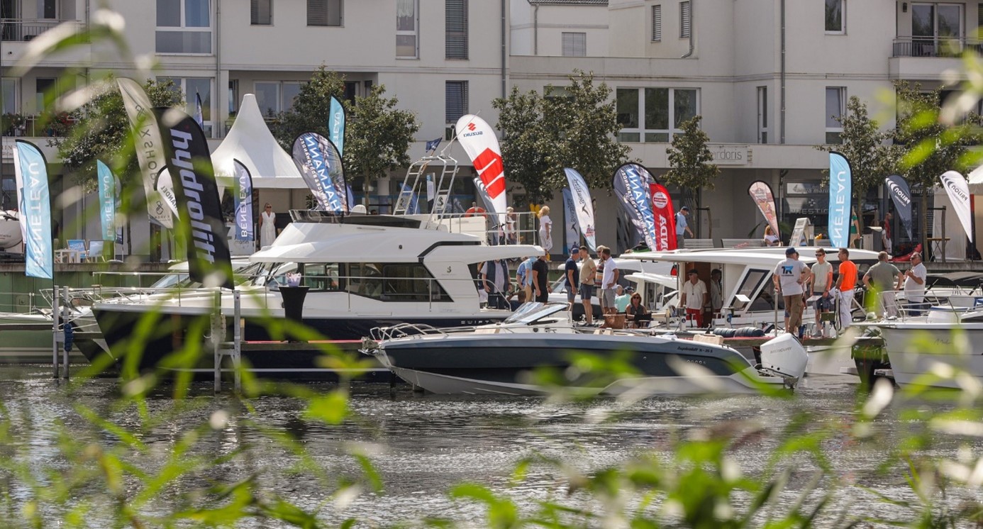 In der Marina in den Havelauen von Werder liegen Boote aller Segmente und Größenklassen an den Stegen. Auf der Promenade im Hintergrund informieren sich Besucher:innen an Verkaufsständen.